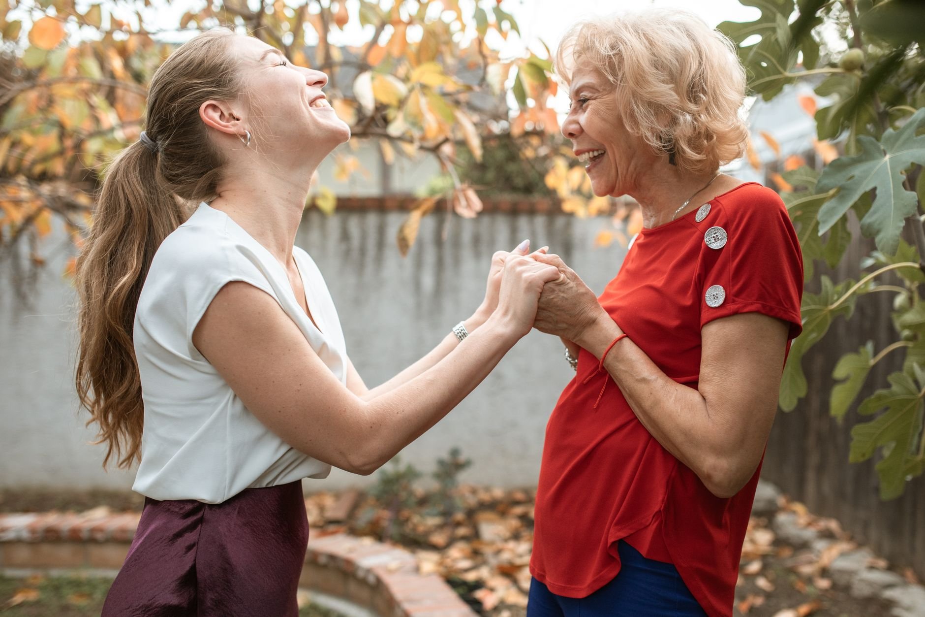 A joyful exchange between two women, young and senior adult, holding hands and laughing outdoors.